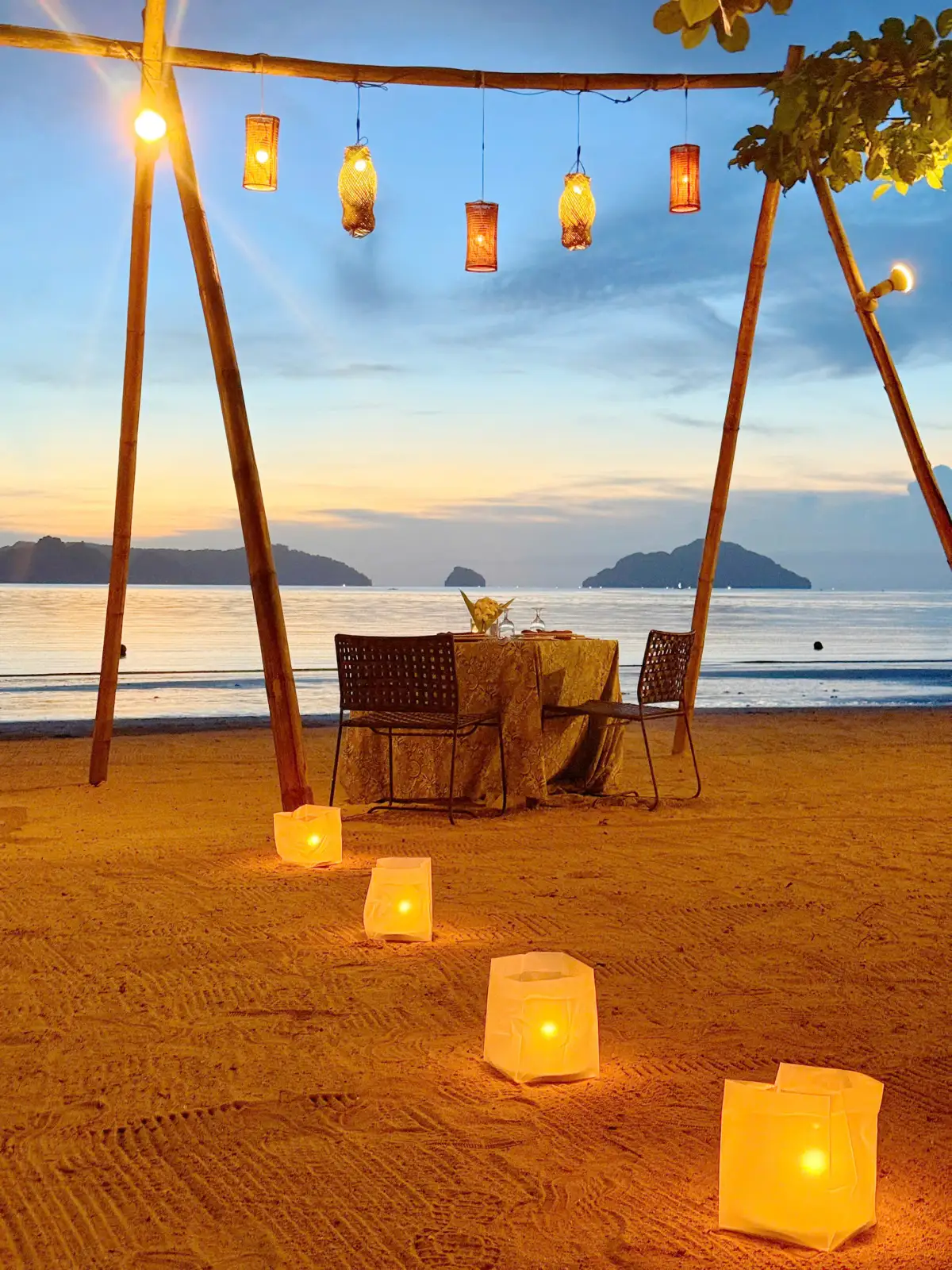 Bàn ăn tối cho hai người trên bãi cát, đèn lồng treo, phía xa là các đảo lúc hoàng hôn