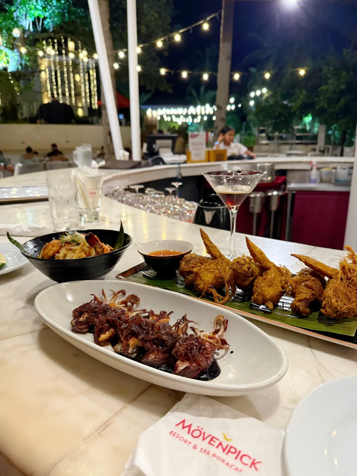Mực nướng, cánh gà chiên và cocktail trên quầy bar đá cẩm thạch, khăn ăn in tên Mövenpick