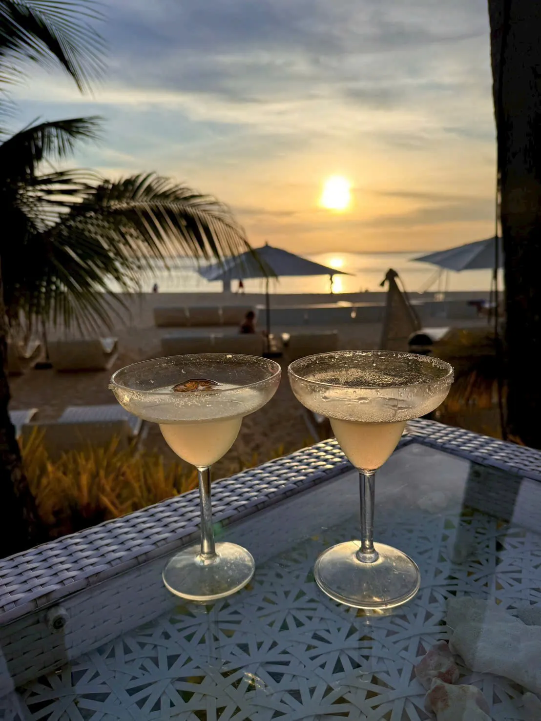 Hai ly cocktail trên bàn, phía sau là hoàng hôn trên biển